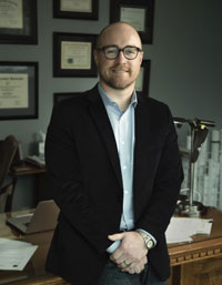 Andrew Carlton Andrew Carlton: Photograph of a man wearing a black blazer, light blue shirt, and wristwatch standing in an office setting. Background includes framed certificates on a wall, a wooden desk with papers, and architectural models or blueprints.